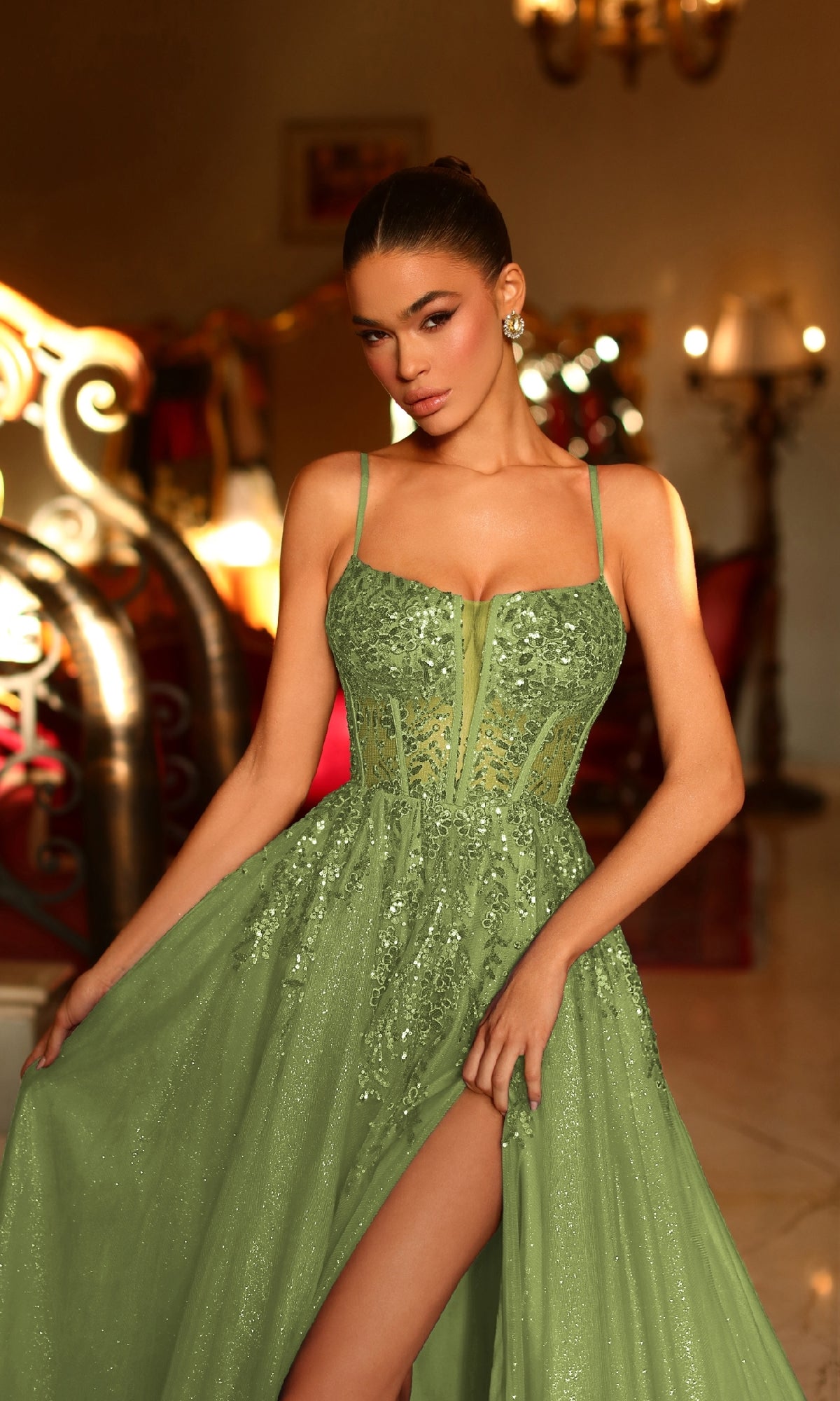 A woman wears the Long Olive Green Prom Ball Gown: Tina Holly TE216, featuring thin straps, a thigh-high slit, and shimmering sequins in an elegant indoor setting.
